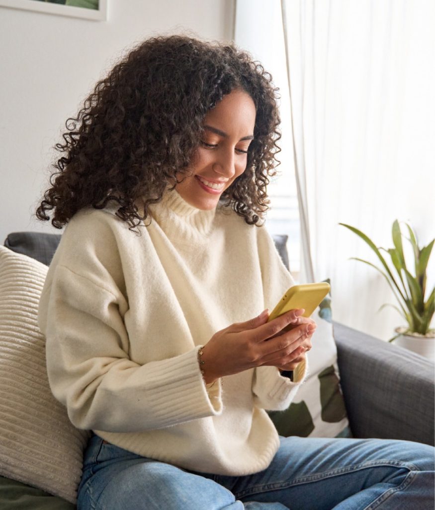 A woman with curly hair sits on a couch, smiling while using a yellow smartphone. A plant and cushions are visible in the background.