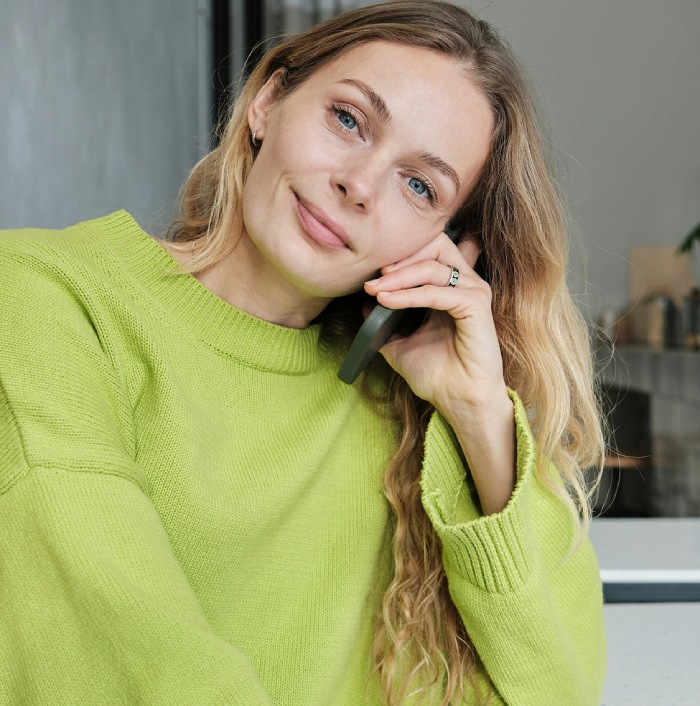 A woman with long blonde hair wearing a green sweater is sitting indoors and holding a phone to her ear, looking at the camera with a slight smile.