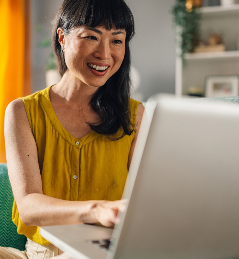 A woman in a yellow sleeveless top sits indoors, smiling while working on a laptop.