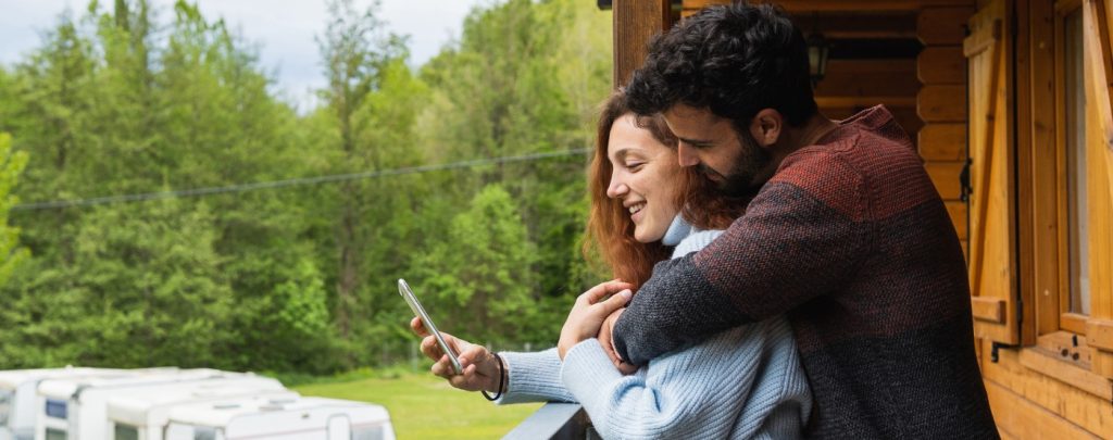 A couple stands on a wooden balcony, smiling and looking at a smartphone together, with trees and parked RVs visible in the background.