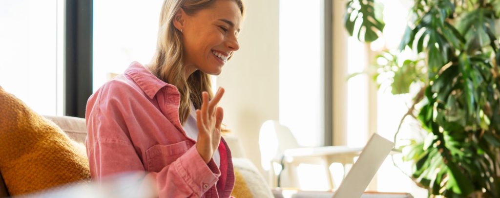 A woman sits on a couch, smiling and waving at a laptop during a video call, with sunlight and plants in the background.