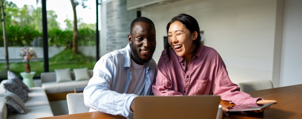 A man and woman sit at a table smiling and looking at a laptop in a bright, modern living room with large windows.