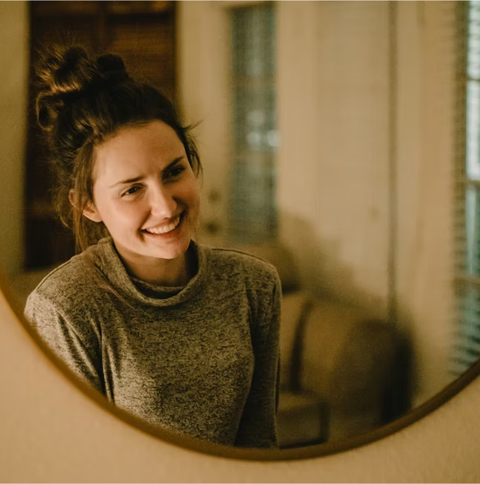 A woman with a relaxed hairstyle and a gray sweater smiles while looking at herself in a round wall mirror in a warmly lit room.