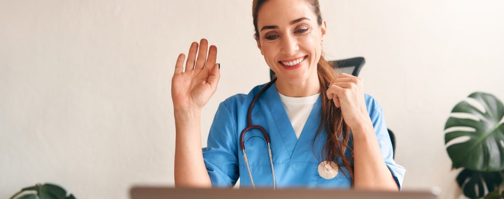 A healthcare professional in blue scrubs with a stethoscope smiles and waves while looking at a laptop, suggesting a virtual consultation.