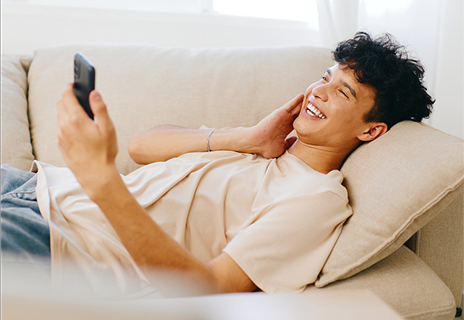 A young man lies on a beige couch, smiling and looking at his smartphone, with his hand resting on his neck.