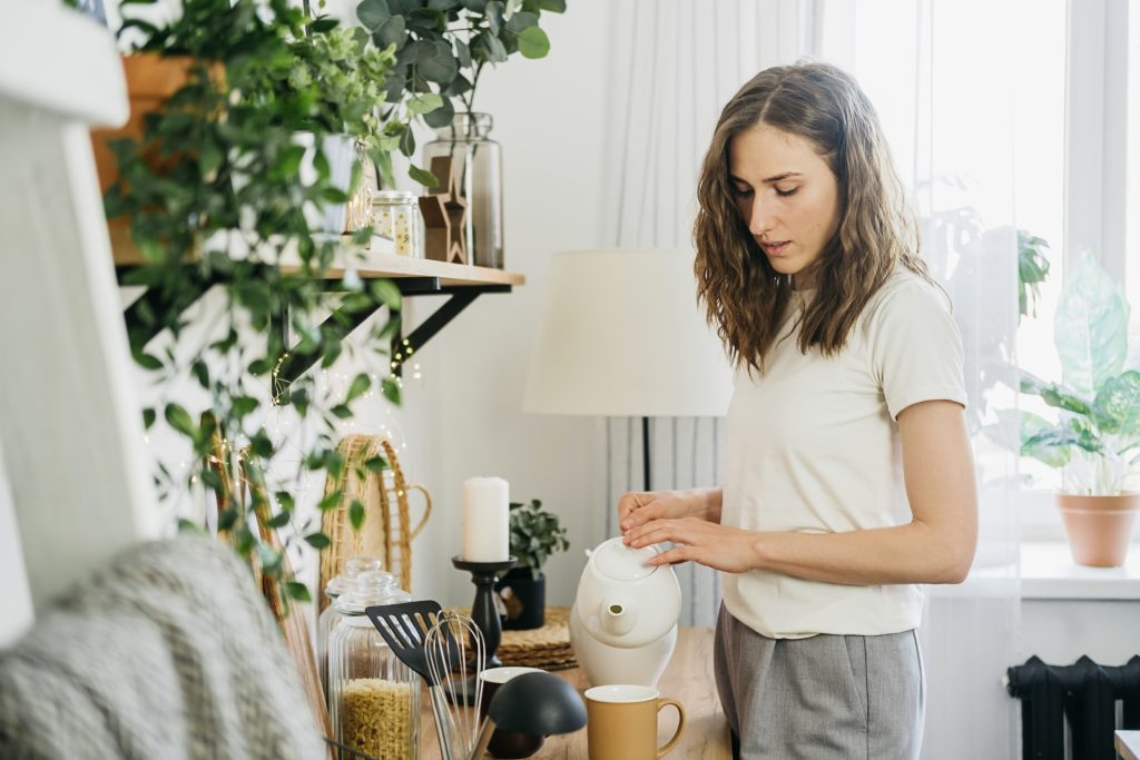 A woman stands in a kitchen by a counter, pouring hot water from a white kettle into a mug, surrounded by plants and kitchen items—a quiet moment of mindfulness and intentional choices.